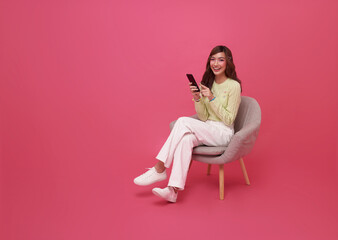 Full length beautiful Asian teen woman using smartphone and sitting on chair isolated on pink...