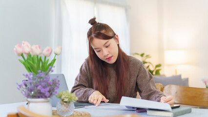 Asian business woman in sweater reading to checking financial document and researching business information on laptop during analyzing about marketing strategy of new startup while working from home