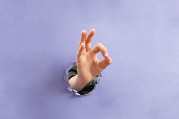 Child hand showing ok sign through a ripped hole in purple color paper background, soft focus close...