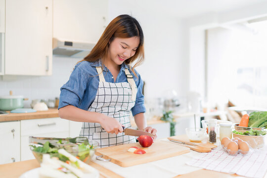 Young asian woman use knife to cutting slices apple on cutting board while preparing fresh vegetables salad in bowl and cooking healthy breakfast food in modern kitchen with healthy lifestyle at home