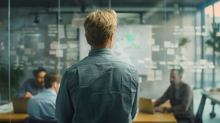 Confident male professional in casual shirt standing with his back to the camera, looking at diverse group of colleagues brainstorming ideas and worki
