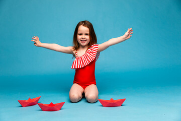 A happy little funny girl in red swimsuit playing with paper boats on blue background. A little...