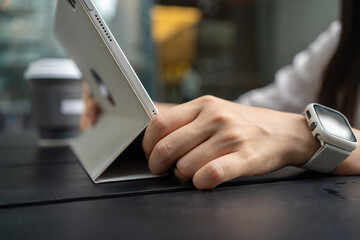 Close up hand of woman wear a white smart watch using digital tablet at coffee shop in urban city.