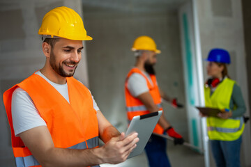 Construction site manager standing  wearing safety vest and helmet, holding a tablet.