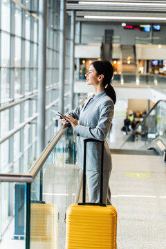 Asian girl in business suit at airport terminal with yellow suitcase looking in window. Business woman with valise looking in panorama window while waiting on flight boarding for vacation.