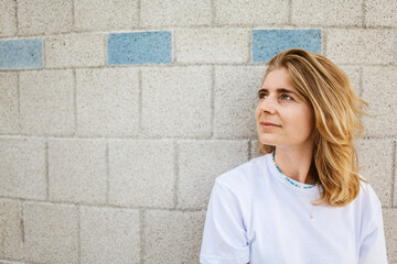 Blond-haired woman in casual white T-shirt looking thoughtful against a brick wall with blue...