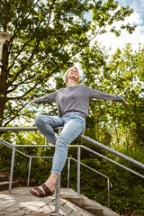 Happy young woman sitting on a railing in a lush green park, enjoying a sunny day with arms outstretched.