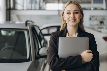Pensive stylish businesswoman manager in suit holding laptop computer, standing near car at car dealership