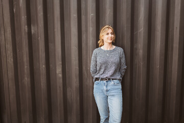 A woman in casual attire stands against a brown corrugated wall, exuding confidence and ease....