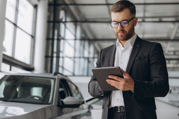 Bearded friendly car seller in glasses and suit standing in car salon and holding tablet