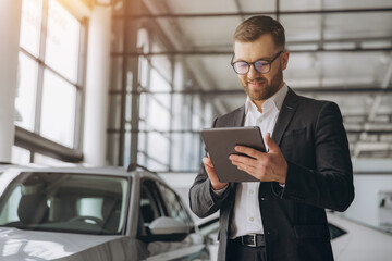 Bearded friendly car seller in glasses and suit standing in car salon and holding tablet