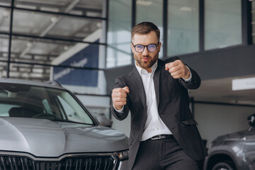 Yes, that's my new car! Customer in car dealership. Happy bearded man new car owner shows driving a...