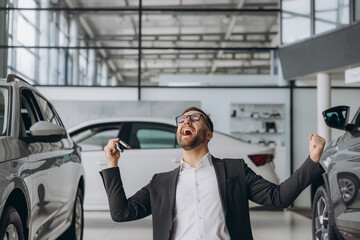Yes, that's my new car! Customer in car dealership. Young happy bearded man knelt down with keys in hand