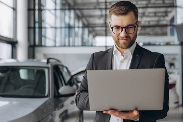 Modern bearded man in glasses and suit vehicle sales consultant using laptop inside car dealership