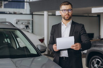 Handsome bearded car dealership worker in suit is holding a folder and smiling while standing near...