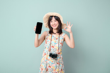 Travel promotion screen concept. Happy Asian woman wearing casual dress and hat with camera showing mobile phone screen isolated on pastel green background.
