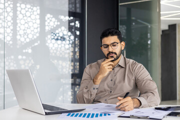 Focused man working on laptop in modern office, studying paperwork and analyzing data. Concentration and dedication to his job. Professional and serious businessman in corporate workplace