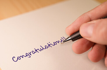 Close-up image of a hand holding a pen, writing the word Congratulations on a sheet of paper....