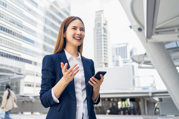 Smiling asian businesswoman holding smartphone while standing in front of modern business building,...
