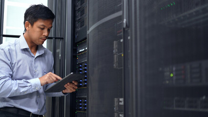 A technician in a blue shirt uses a tablet while inspecting a server rack in a modern data center,...