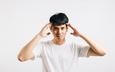 A portrait of an Asian young man with a sad, tired expression, holding his head in his hands due to stress and a painful headache. Studio shot isolated on a white background.