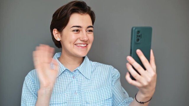 Positive telephone conversation, cheerful young woman using a video call on a smartphone waves her hand in a welcoming gesture and says Hello to her interlocutor