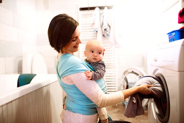 Mother holding small baby while loading the washing machine with dirty laundry, carrying him around...