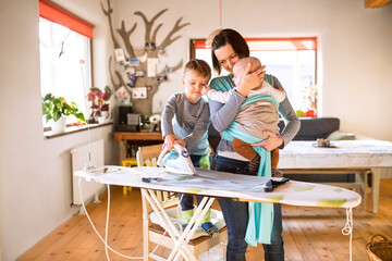 Mother carrying small baby in wrap, older son helping with ironing. Family doing household chores.