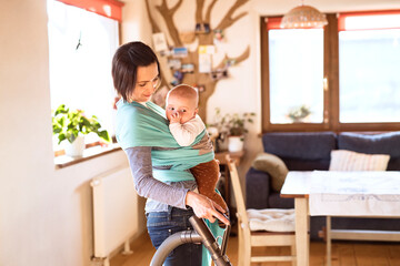 Mother holding small baby while vacuuming floor, carrying him around house in baby wrap. Household...