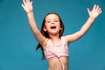 Portrait of a little excited smiling girl raising her hands in joy and looking away on a blue...