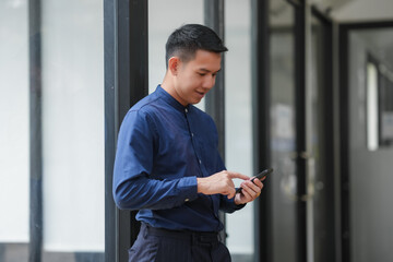 Businessman in blue shirt using smartphone against modern office background, engaging with technology for communication.