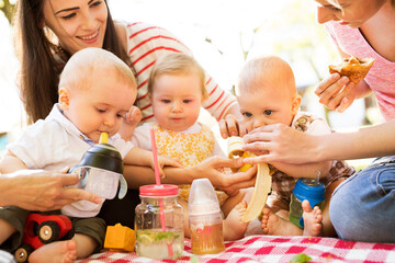 Mothers and babies enjoying group picnic outdoor in park, sitting on picnic blanket and preparing food and drinks.