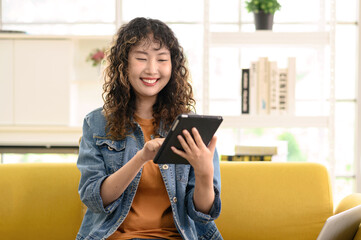 Asian woman sitting on cozy sofa reading or working with tablet. Lifestyle image of beautiful young...