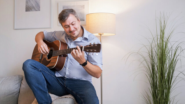 Musical hobby. Guitar skill. Happy smiling man sitting on sofa playing string instrument enjoying learning chords in living room interior at home copy space.