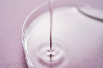 Serum or cosmetic oil flows into a transparent bowl on a purple background.