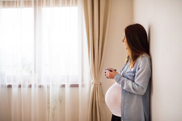 Pregnant woman leaning against wall in her home, drinking herbal tea. Red raspberry leaf tea to...