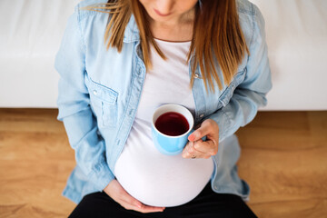 Pregnant woman sitting on floor in front of sofa, drinking herbal tea. Red raspberry leaf tea to...