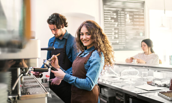 Two young baristas working in coffee shop, standing by counter. University students working part-time in cafe.
