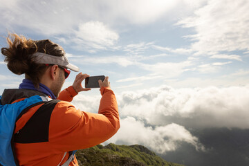 A man wearing an orange jacket is taking a picture of the clouds