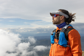 A man in an orange jacket stands on a mountain top, looking out over the clouds