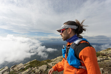 A man wearing an orange jacket and sunglasses is standing on a mountain top