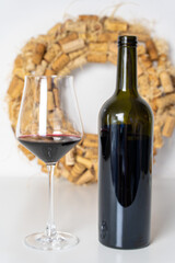 A glass of wine with a wine bottle on a white table, a wreath of wine corks in the background. Selected focus.