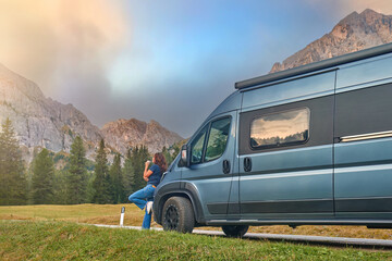 Woman leaning against a camper van, enjoying a scenic mountain view with rocky peaks and pine trees...