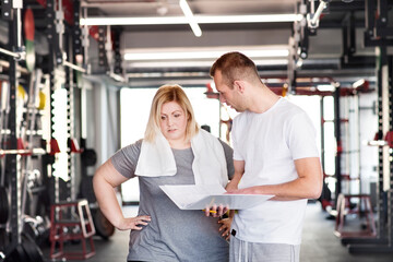 Overweight woman consulting with trainer, discussing daily workout plan. Personal trainer talking with client, discussing exercise program in gym.