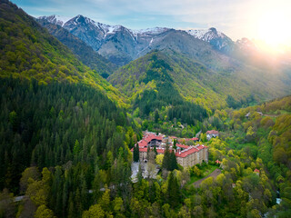 Aerial view of Rila Monastery nestled in the lush green forests of Rila Mountain with snow-capped peaks in the background, Bulgaria. The historical complex features red-roofed buildings.
