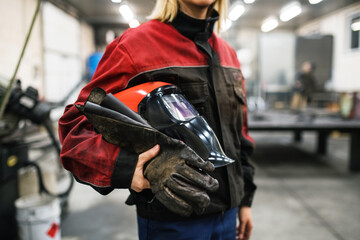 Close up of welding mask in hands of woman welder in workshop. Female worker operating welding machine, wearing protective clothing and a welding mask.