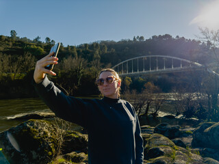 Woman Taking Selfie by River with Bridge in Background