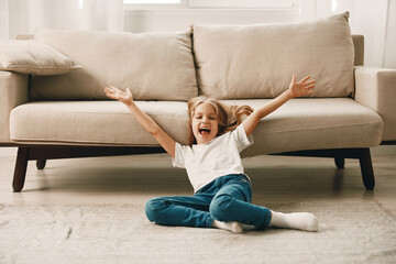 Little girl sitting on floor with arms outstretched, beige couch in background, cozy home interior...