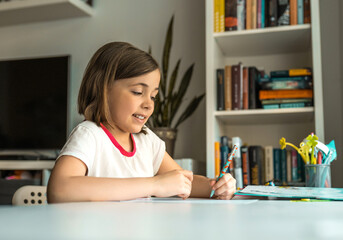 Little girl wries something with left hand at a desk in front of a book shelves. Children education concept.