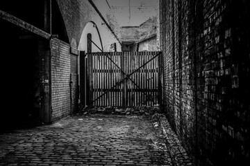 Black and white photo of deserted cobblestone alley with industrial gate and brick walls, creating...